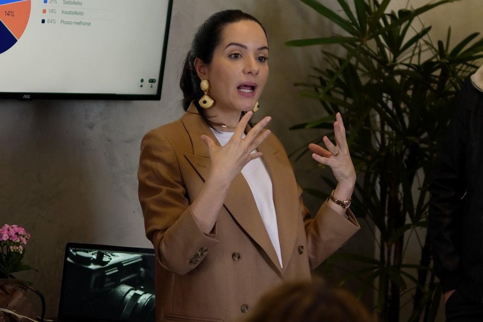 Erika Furtado de blazer camel falando para uma plateia, com um gráfico projetado na tela ao fundo