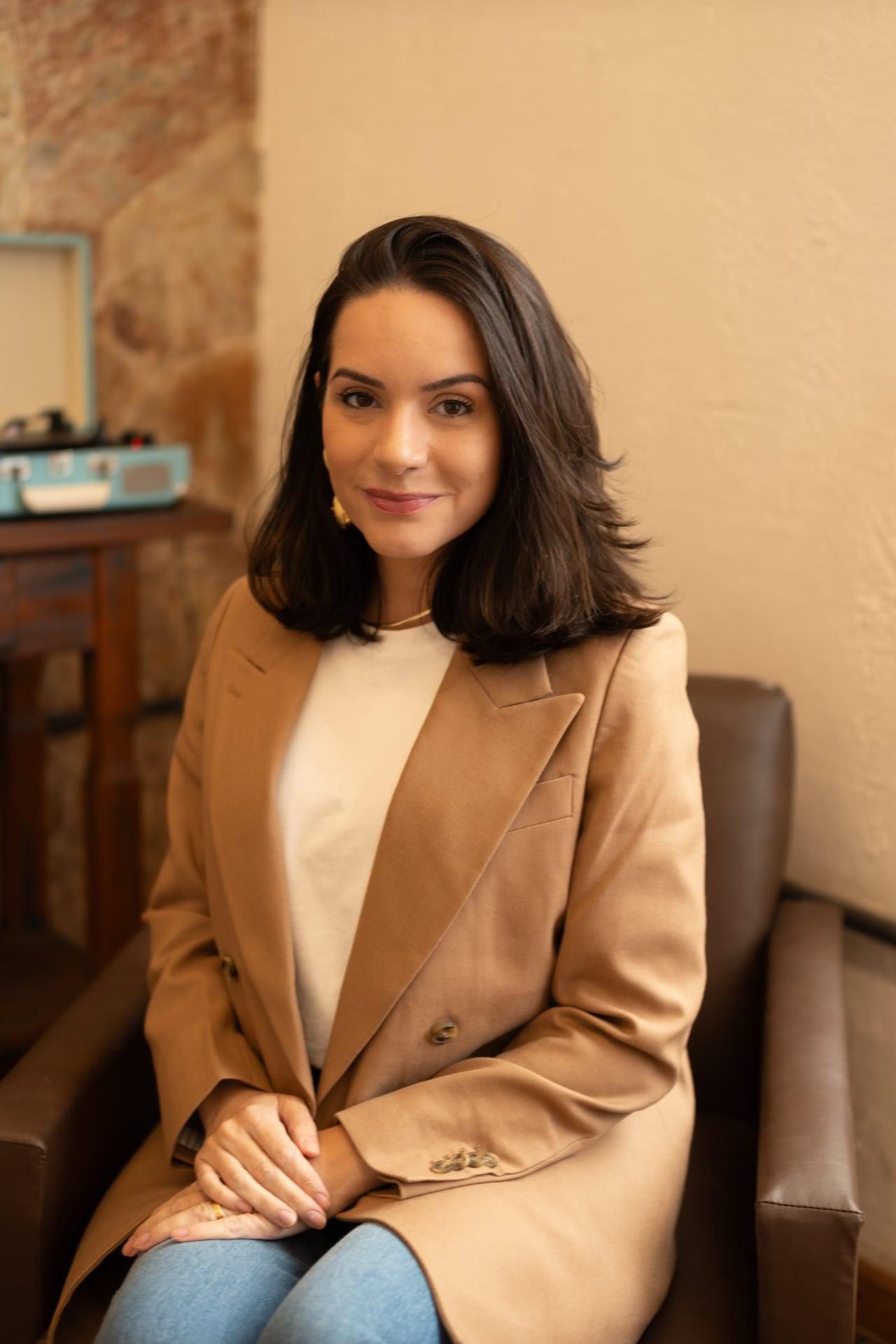 Erika Furtado de blazer camel, sentada numa poltrona de couro, sorrindo para a câmera